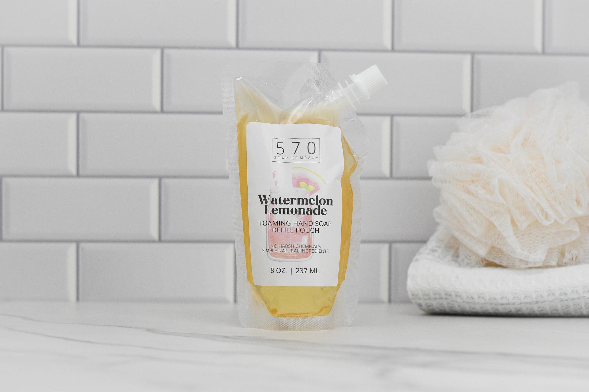 A pouch of Watermelon Lemonade Scented Foaming Hand Soap from 570 Soap Co. is displayed on a marble countertop, set against a white subway tile backsplash. It sits next to a stack of folded white towels and a beige bath sponge, providing moisturizing benefits with every use.