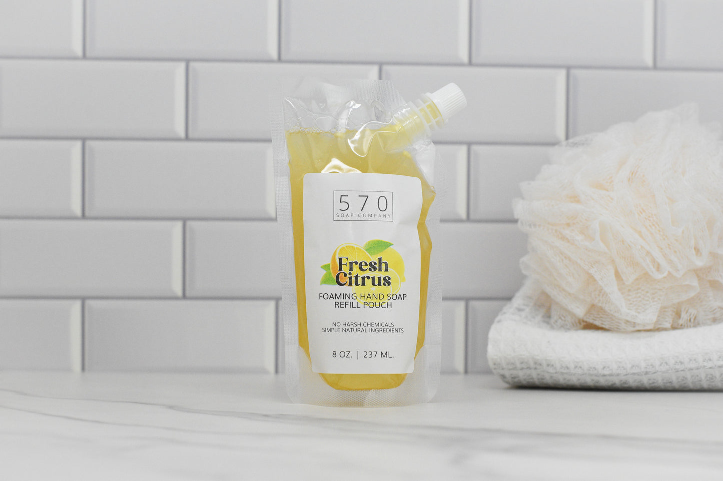 A refill pouch of 570 Soap Co.s Fresh Citrus All Natural Foaming Hand Soap is on a bathroom counter with white tiles. A folded white towel and bath sponge nearby enhance its fresh citrus scent with essential oils.