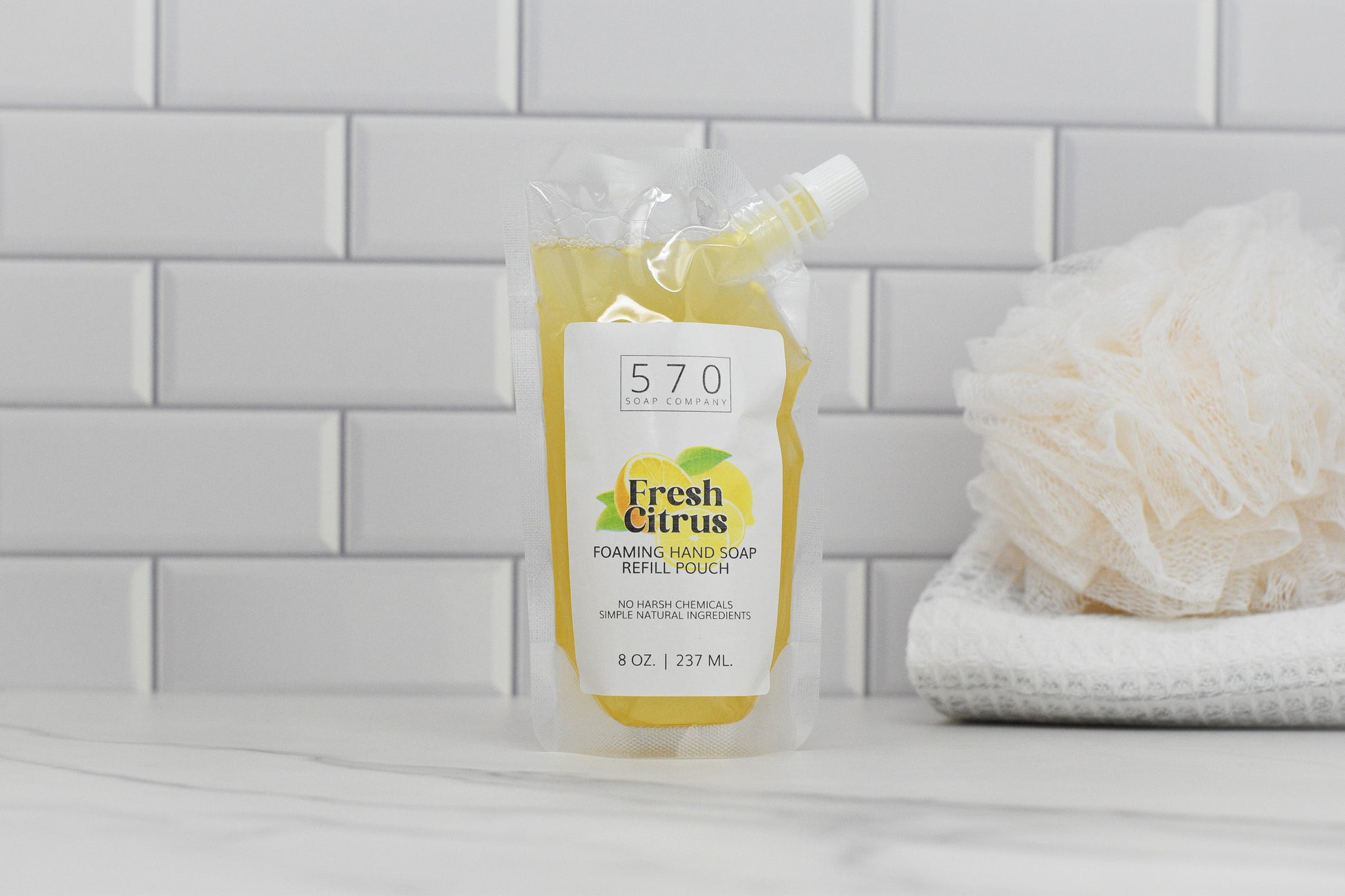 A refill pouch of 570 Soap Co.s Fresh Citrus All Natural Foaming Hand Soap is on a bathroom counter with white tiles. A folded white towel and bath sponge nearby enhance its fresh citrus scent with essential oils.