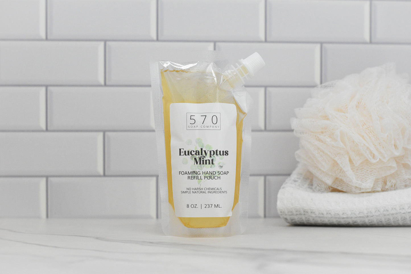 A refill pouch of Eucalyptus Mint All Natural Foaming Hand Soap by 570 Soap Co. is on a bathroom counter. The transparent pouch shows the brand, while soft, neutral-colored tiles and a folded white loofah in the background create a serene and refreshing ambiance.