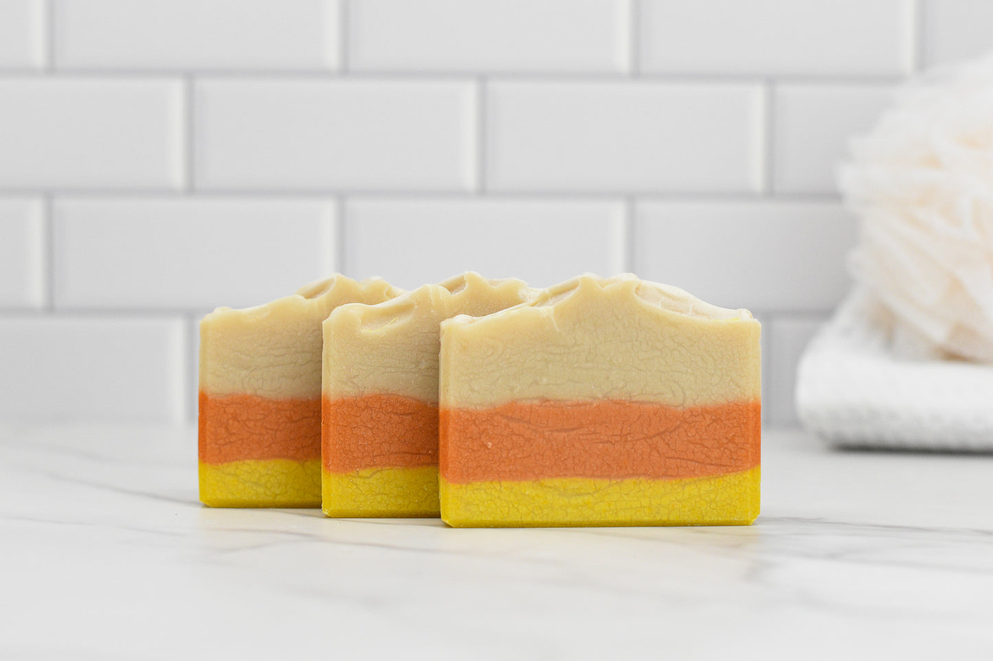 Three bars of 570 Soap Co.'s Candy Corn Bar Soap - Coconut Milk, in hues of cream, orange, and yellow, are neatly placed on a white marble surface. A white tiled wall and a beige bath sponge in the background enhance the minimalist bathroom vibe while emphasizing the natural ingredients used for a gentle cleanse.