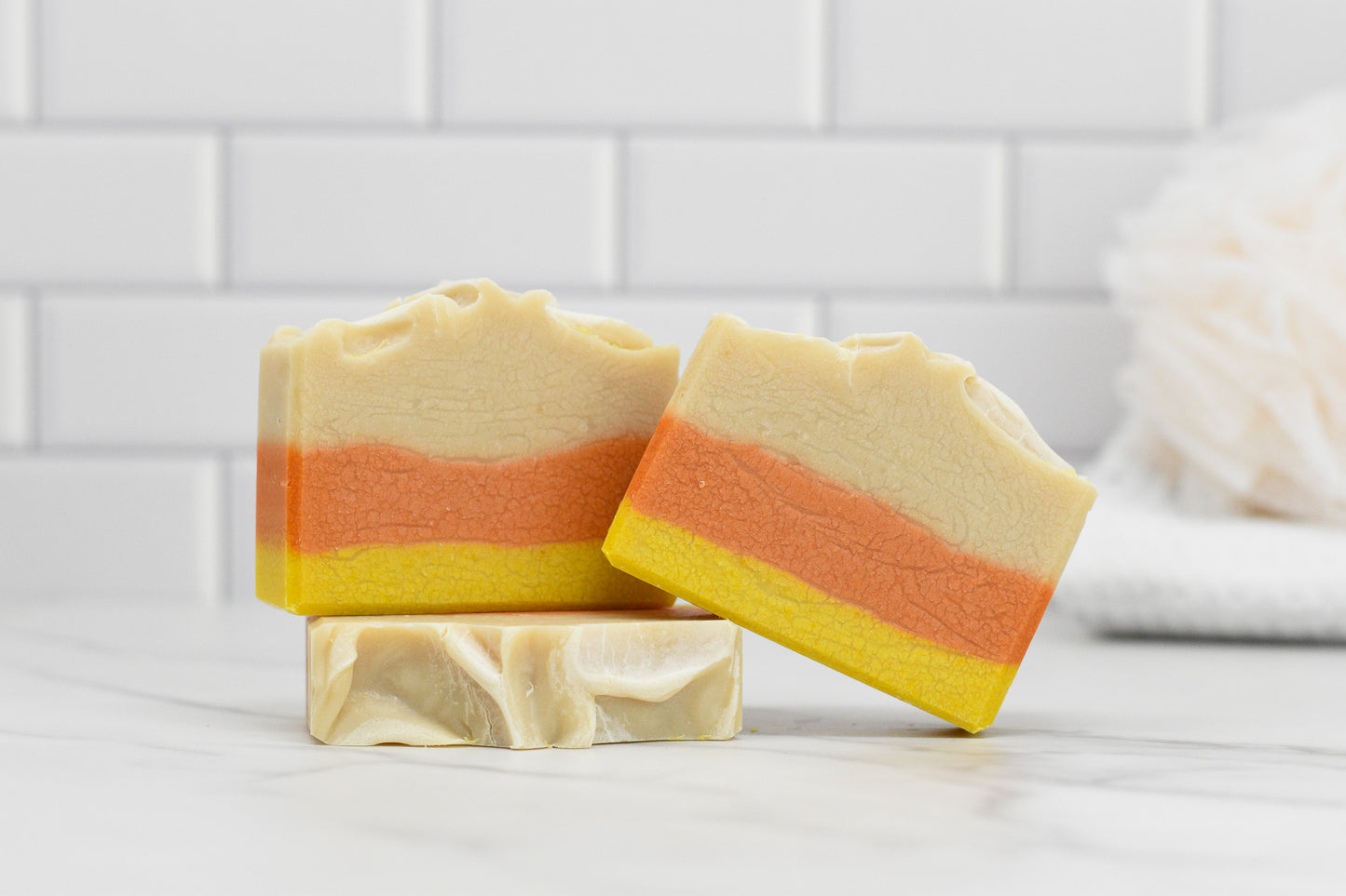 Three bars of the Candy Corn Bar Soap - Coconut Milk from 570 Soap Co. are elegantly displayed on a marble surface, highlighting layers of cream, orange, and yellow. Made with natural ingredients for a gentle cleanse, their beauty is enhanced by a white background and a soft bath accessory nearby.