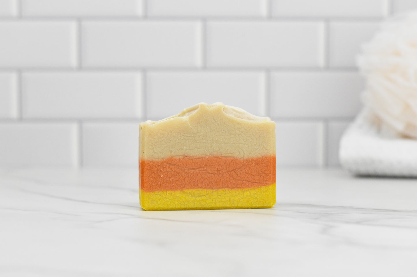 The Candy Corn Bar Soap - Coconut Milk by 570 Soap Co. showcases three layers in cream, orange, and yellow, resting on a marble surface and providing a gentle cleanse. The white tiled wall and a white loofah enhance the background ambiance.