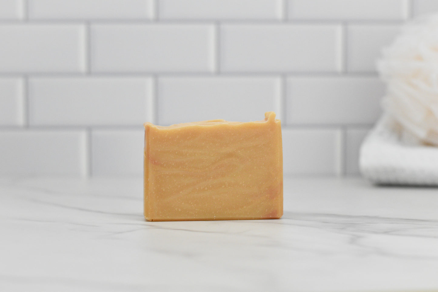 A bar of 570 Soap Co.'s Fall Spice Bar Soap rests on a marble surface, infused with Autumn Aromas. The light orange hue and gentle swirls capture the essence of fall. In the background, a white tiled wall with a blurred bath sponge enhances this cozy seasonal scene.