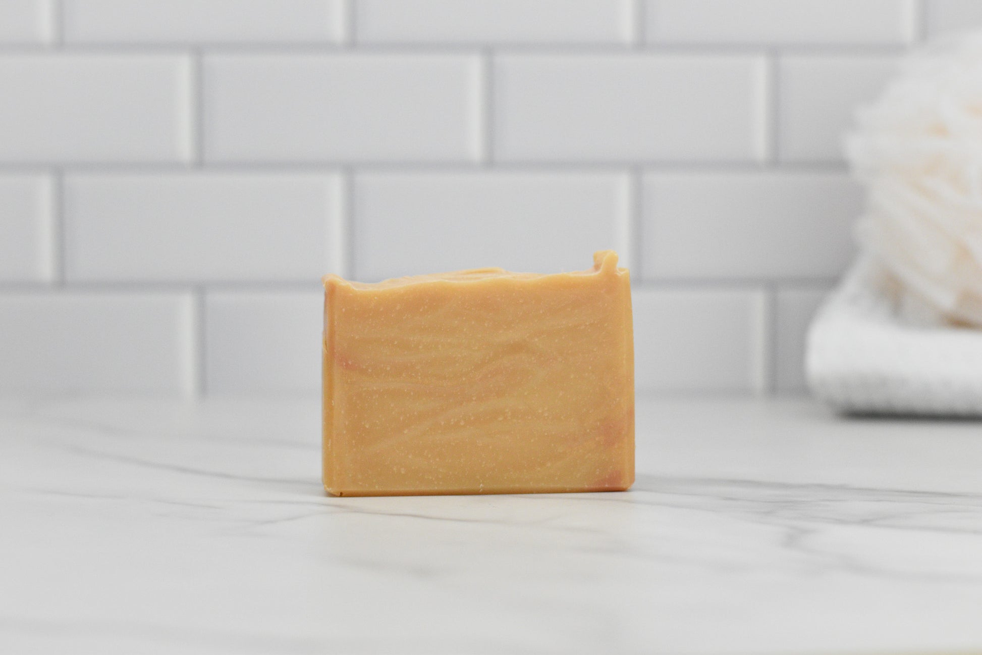 A bar of 570 Soap Co.'s Fall Spice Bar Soap rests on a marble surface, infused with Autumn Aromas. The light orange hue and gentle swirls capture the essence of fall. In the background, a white tiled wall with a blurred bath sponge enhances this cozy seasonal scene.