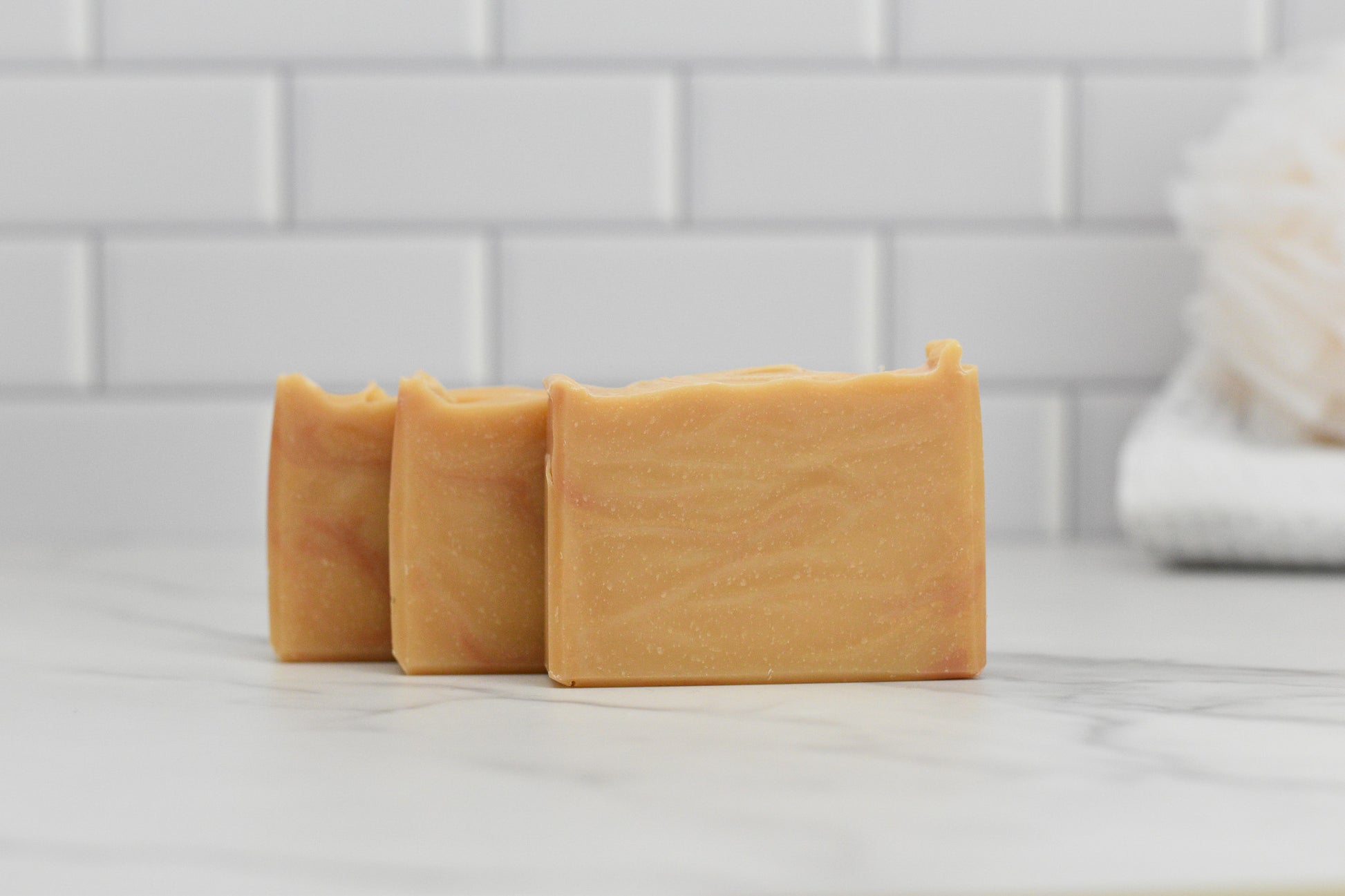 Three bars of 570 Soap Co.'s Fall Spice Bar Soap, featuring a marbled tan and orange design, are elegantly placed on a white marble countertop. The scene is complemented by white subway tiles in the background alongside a fluffy bath sponge, capturing the essence of cozy Autumn Aromas.