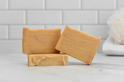 Three bars of Fall Spice Bar Soap by 570 Soap Co., with their beige hue, are stacked on a white marble surface against a backdrop of white subway tiles. The seasonal scent gently permeates the air, enhancing the cozy ambiance while a white bath sponge sits invitingly in the corner.