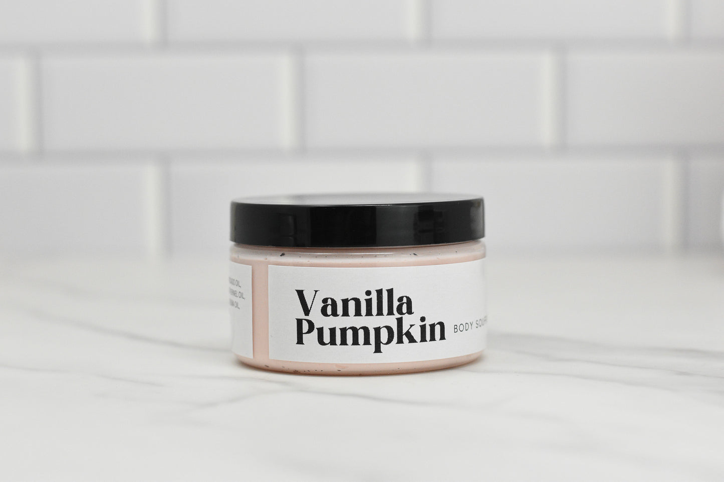 A jar of the Vanilla Pumpkin Body Butter - SEASONAL by 570 Soap Co., featuring a black lid, sits on a light-colored surface beside its companion body scrub. The white label with black text is set against crisp white subway tiles, offering warmth and hydration.