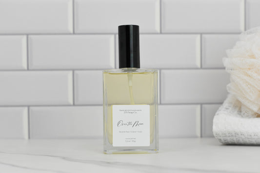 A rectangular glass bottle with a black cap, containing a light yellow liquid, sits on a white marble surface. The label reads "Over the Moon Eau de Parfum" by 570 Soap Co., indicating its luxurious fragrance. In the background, there's a soft, folded towel against white subway tiles.