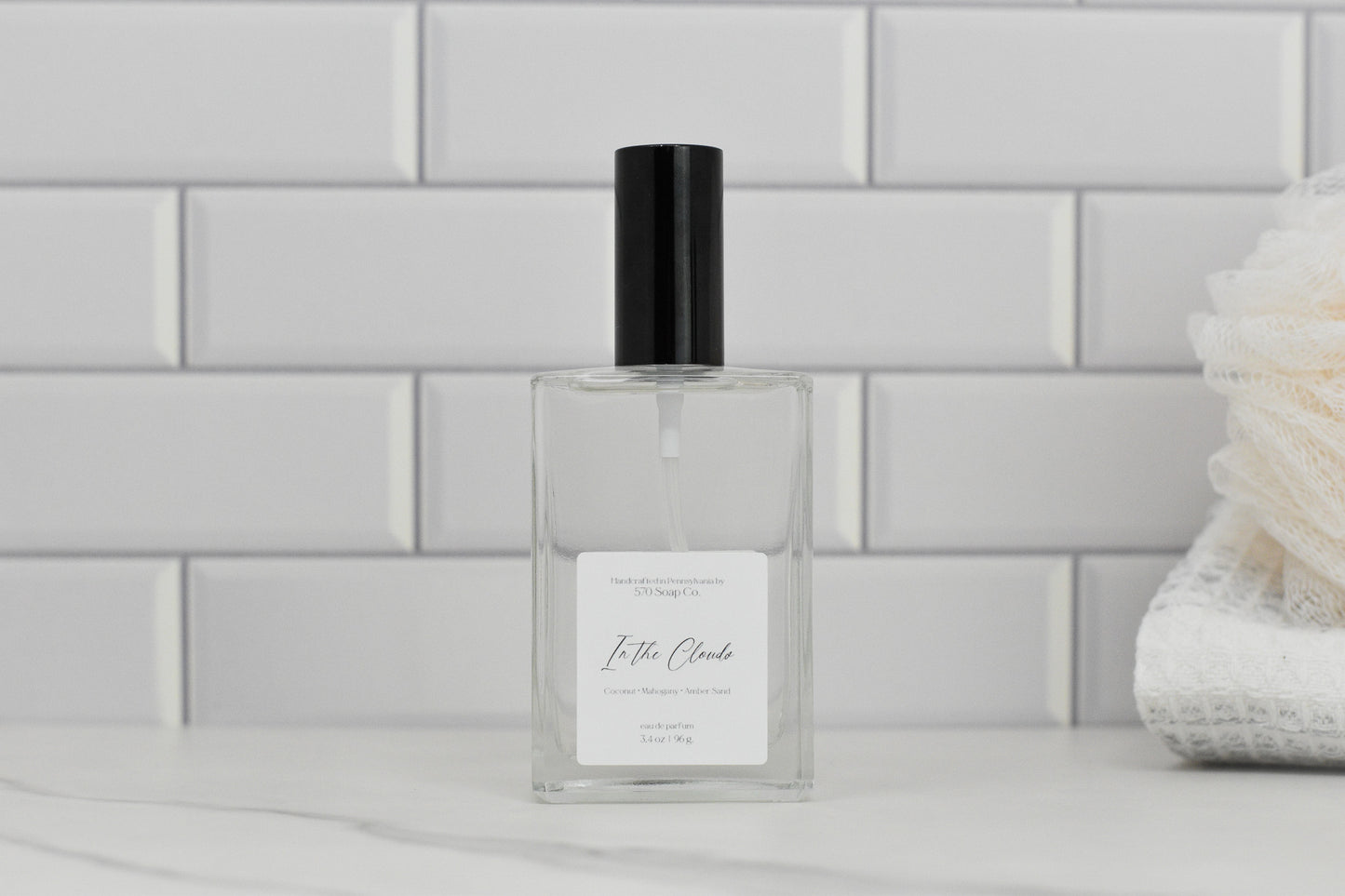 A clear rectangular glass bottle with a black cap rests on a marble surface, exuding an air of luxury. The label reads "In the Clouds Eau de Parfum" by 570 Soap Co., suggesting a tropical fragrance essence. In the background, a stack of white towels softly fades against white subway tiles.