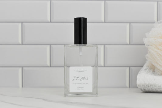 A clear rectangular glass bottle with a black cap rests on a marble surface, exuding an air of luxury. The label reads "In the Clouds Eau de Parfum" by 570 Soap Co., suggesting a tropical fragrance essence. In the background, a stack of white towels softly fades against white subway tiles.