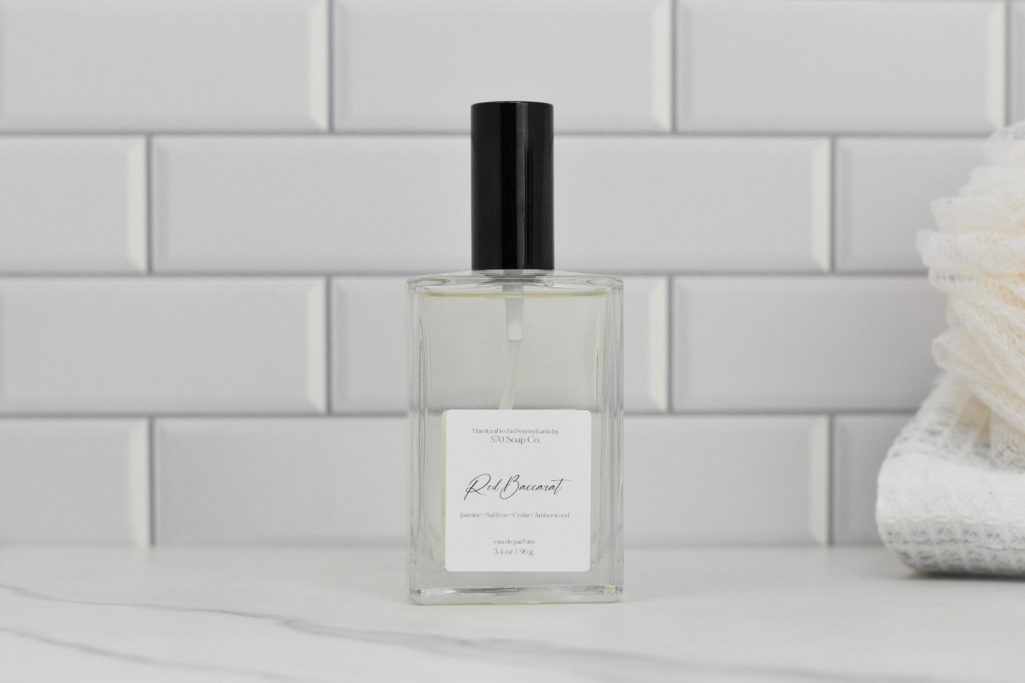 A clear glass perfume bottle with a black cap is set against a backdrop of white subway tiles. The label reads Red Baccarat Eau de Parfum, suggesting a luxurious scent with hints of jasmine, white cedar, and vetiver. To the right, a white bath accessory enhances the elegance associated with 570 Soap Co.'s premium fragrance.