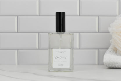 A clear glass perfume bottle with a black cap is set against a backdrop of white subway tiles. The label reads Red Baccarat Eau de Parfum, suggesting a luxurious scent with hints of jasmine, white cedar, and vetiver. To the right, a white bath accessory enhances the elegance associated with 570 Soap Co.'s premium fragrance.