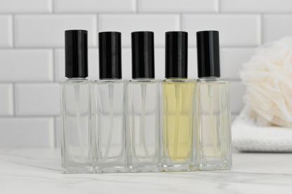 Five glass bottles of In the Clouds Eau de Parfum from 570 Soap Co., each topped with a black cap and filled with varying levels of clear and light yellow fragrance, are lined up on a marble surface. The luxury collection is set against white subway tiles and complemented by a soft, white bath accessory, exuding an air of elegance and style.