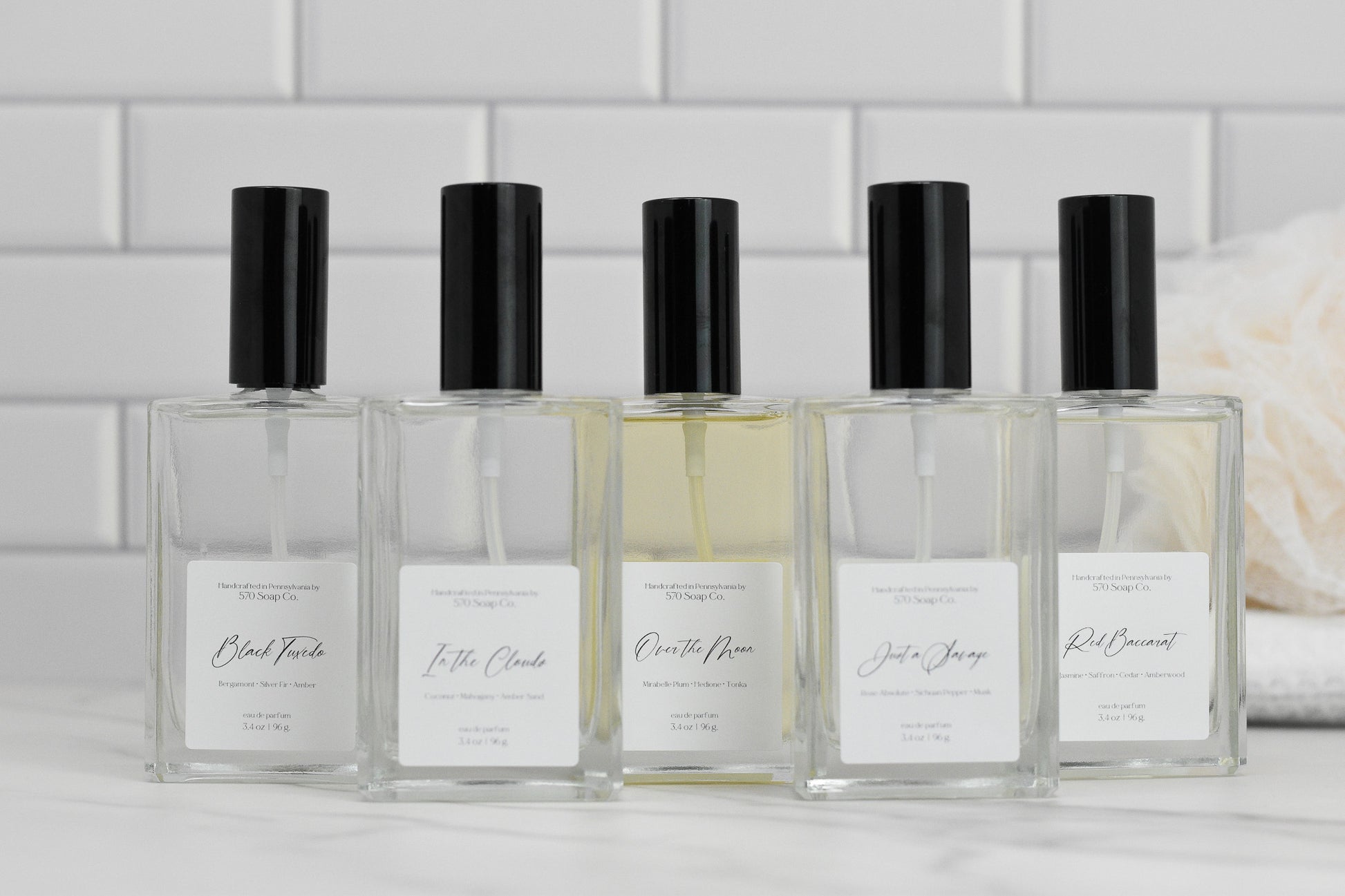 Five rectangular perfume bottles with black caps are lined up against a white tiled background, each prominently displaying a luxury fragrance name. On the right, a beige loofah is partly visible, accentuating the elegance of these Eau de Parfum selections from 570 Soap Co., including the "Over the Moon" fragrance.