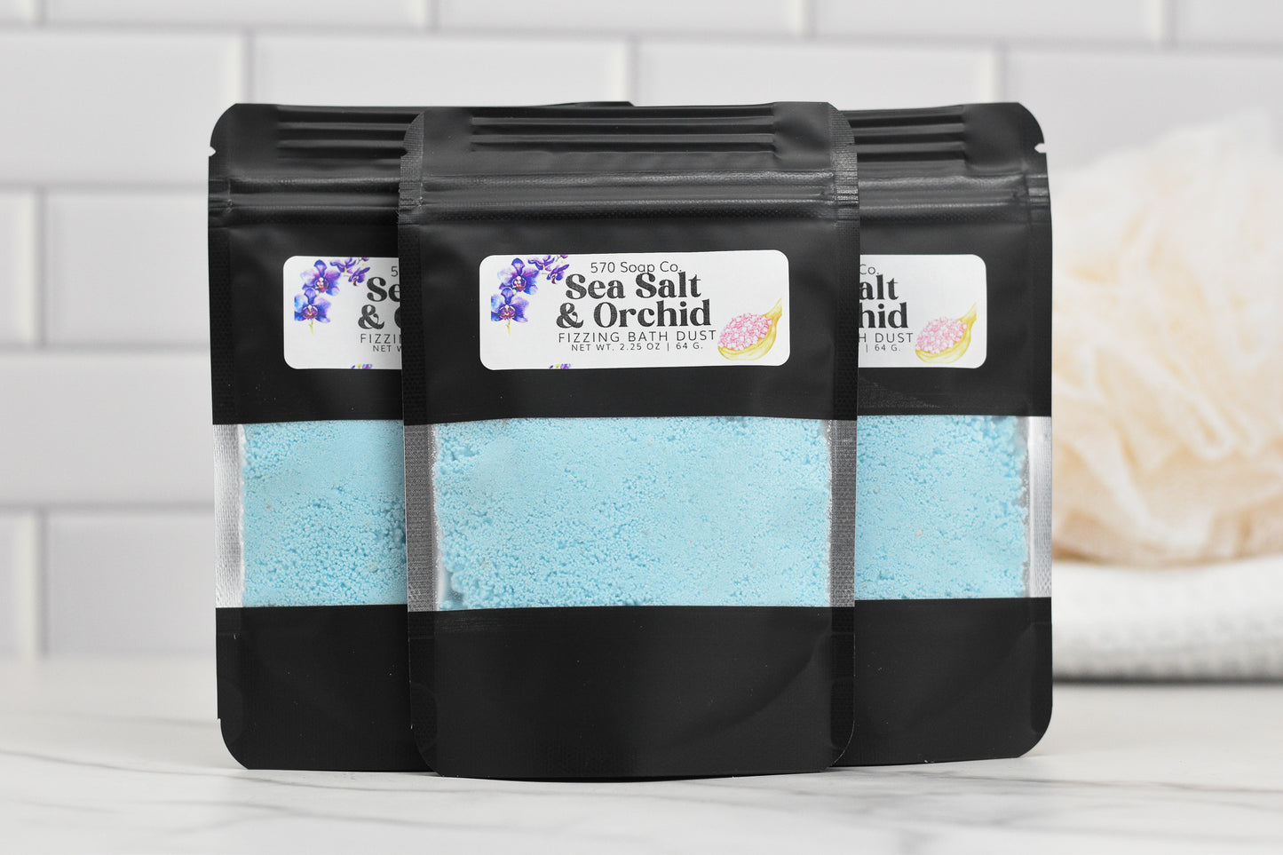 Three black stand-up pouches of Sea Salt & Orchid Fizzing Bath Dust by 570 Soap Co. are arranged in a row. The pouches feature a clear window showcasing the light blue powder inside, set against a bathroom backdrop.