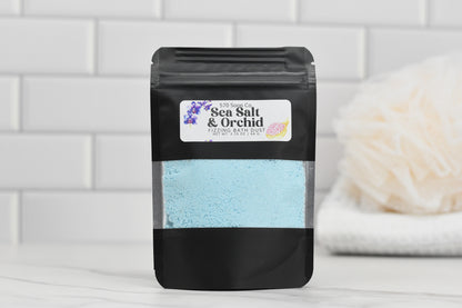 A black package of 570 Soap Co.'s Sea Salt & Orchid Fizzing Bath Dust, containing blue powder, sits on a white surface. In the background, a soft white bath sponge and tiled wall evoke a clean and soothing atmosphere.