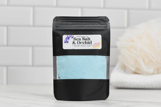 A black package of 570 Soap Co.'s Sea Salt & Orchid Fizzing Bath Dust, containing blue powder, sits on a white surface. In the background, a soft white bath sponge and tiled wall evoke a clean and soothing atmosphere.