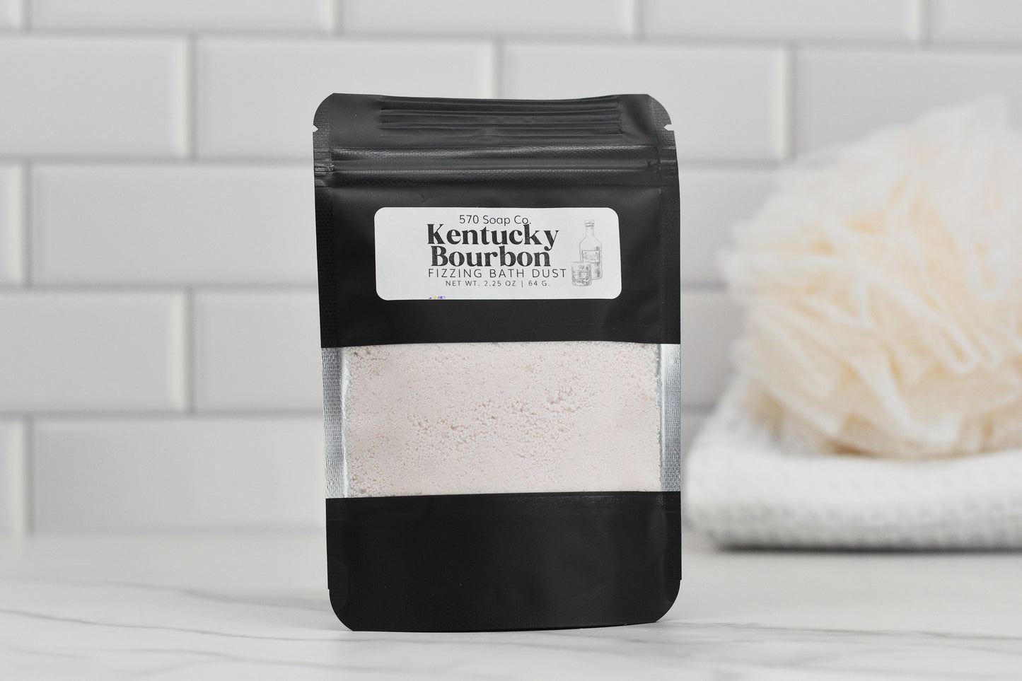 A black pouch bearing the label "Kentucky Bourbon Fizzing Bath Dust" from 570 Soap Co. stands out against a white tiled backdrop. Its partially transparent packaging reveals the fine luxury powder inside, accompanied by a softly blurred bath sponge and towel in the background, suggesting indulgent relaxation.