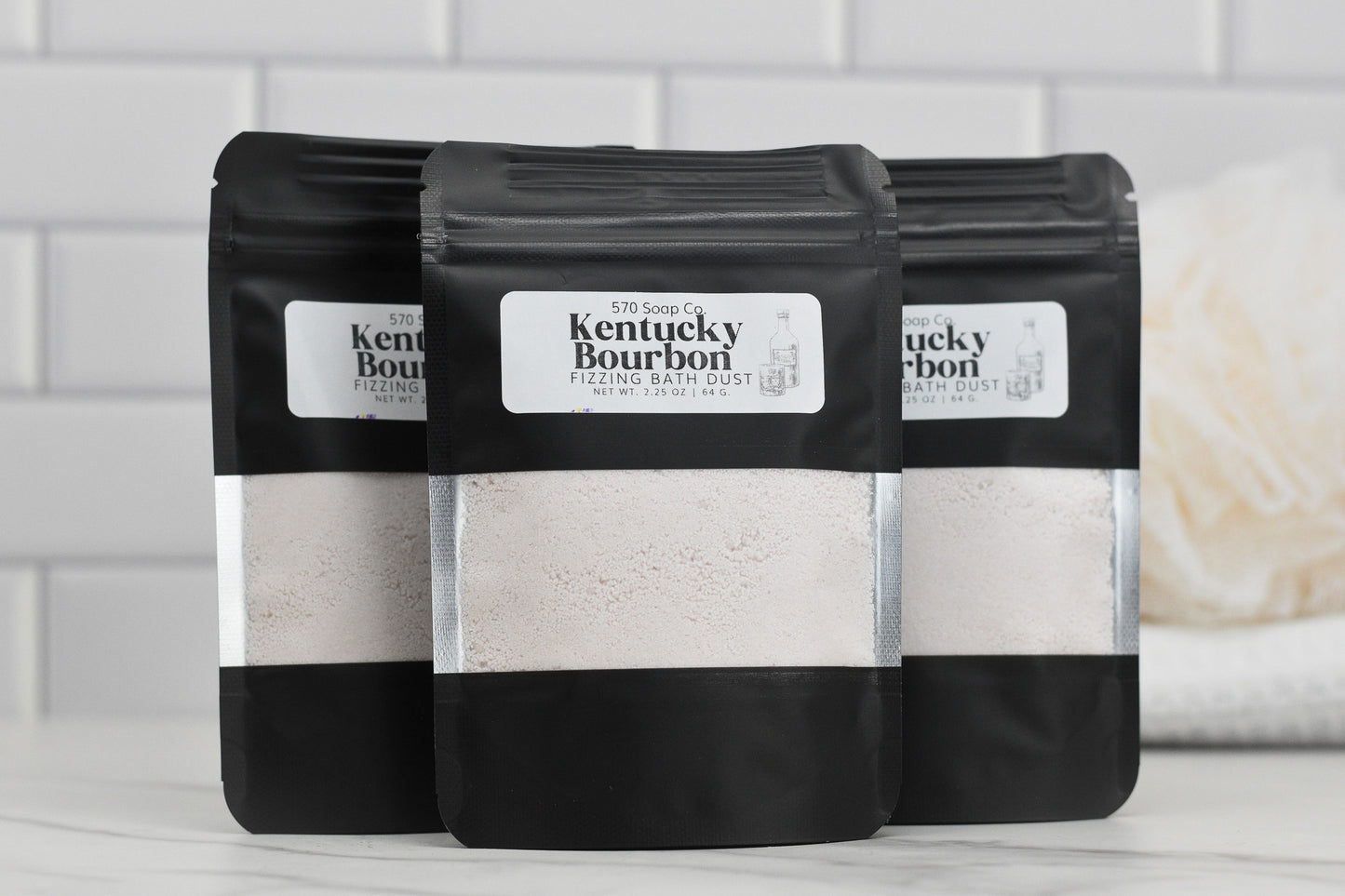 Against a backdrop of white tiles, three black pouches labeled with the name "Kentucky Bourbon Fizzing Bath Dust" from 570 Soap Co. are artfully displayed, showcasing transparent sections filled with sumptuous bath powder for an opulent bathing experience.