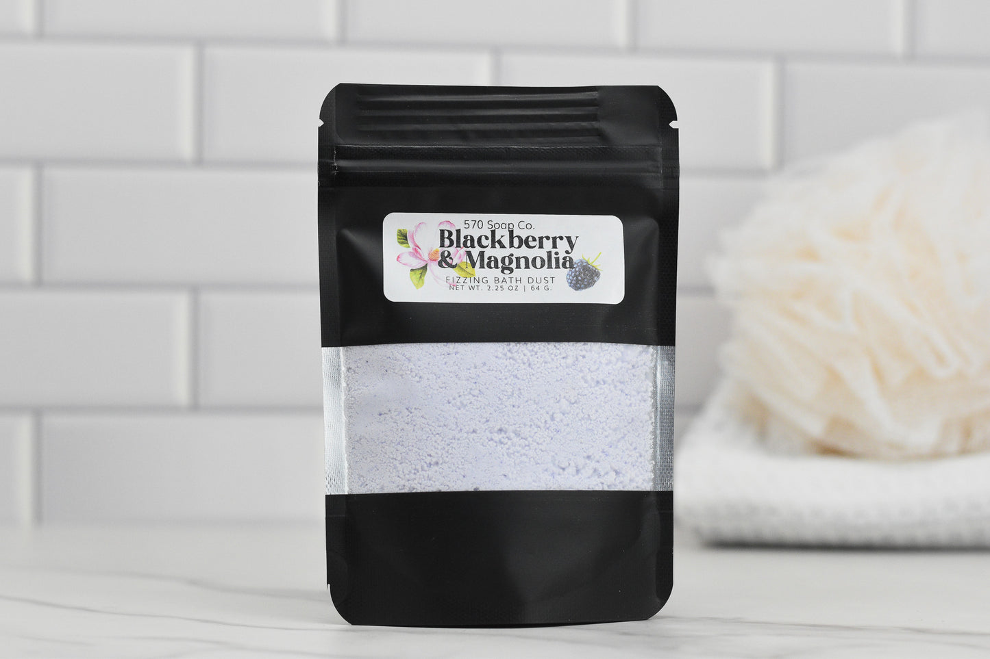 A pouch of 570 Soap Co.s Blackberry & Magnolia Fizzing Bath Dust sits on a white surface in a bathroom, with its label visible against a textured background and complemented by a bath sponge.