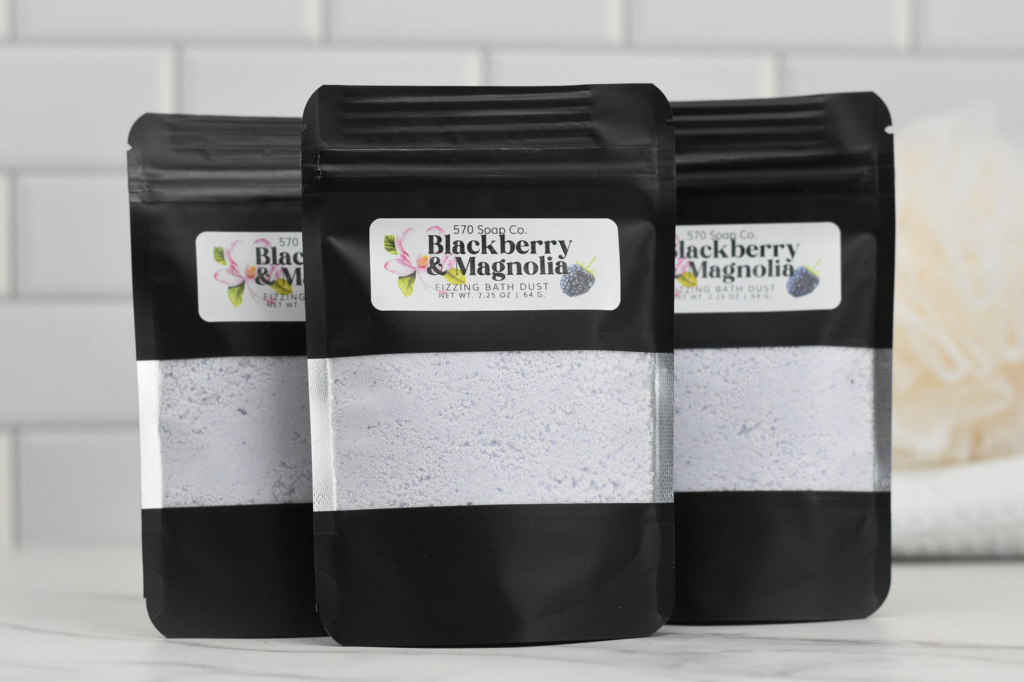 Three black packages of 570 Soap Co.s Blackberry & Magnolia Fizzing Bath Dust are displayed against a white tiled background, featuring floral illustrations on the labels and clear windows showcasing the vibrant bath dust.