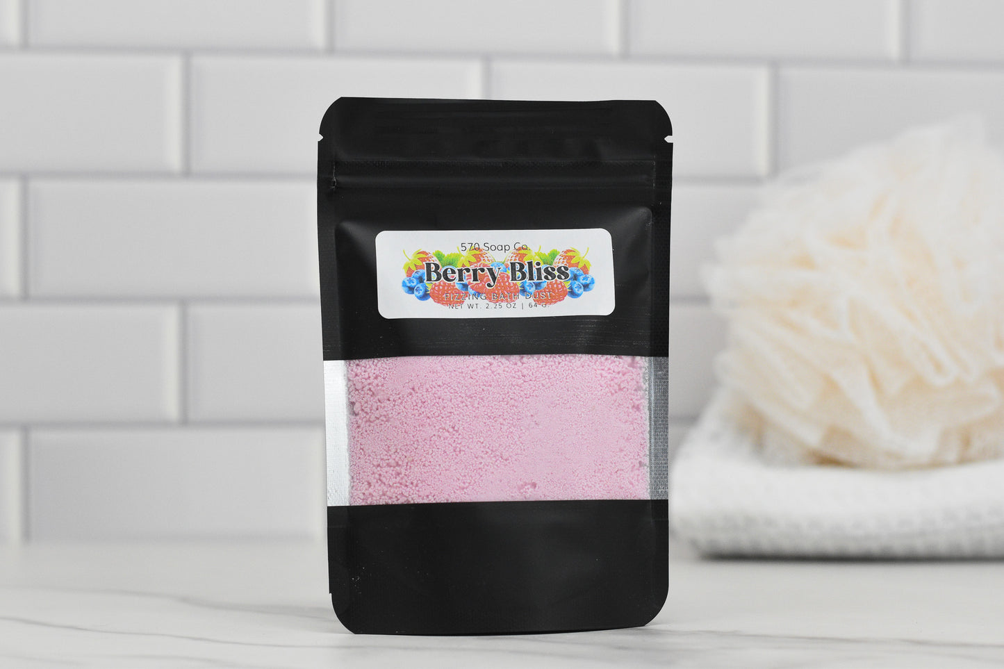 A black pouch from 570 Soap Co. labeled Berry Bliss Fizzing Bath Dust contains pink powder and is placed on a tiled surface. In the blurred background, a white loofah is visible, hinting at a bathroom setting filled with skin-softening ingredients.