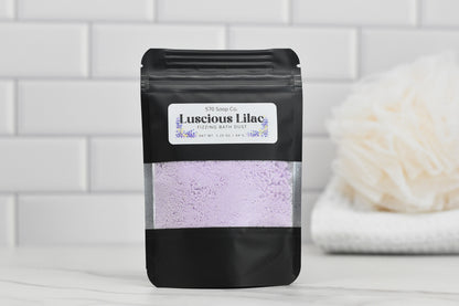 A pouch marked with "Luscious Lilac Fizzing Bath Dust" from 570 Soap Co., containing light purple powder, is displayed on a white surface. The setting includes white subway tiles and a cream-colored bath sponge, inviting you to indulge in effervescent bubbles and lilac dreams.