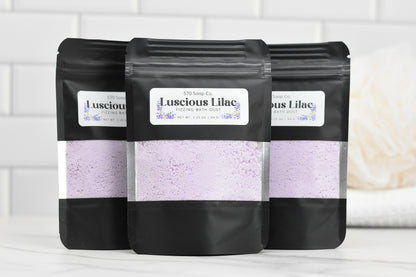 Three black pouches featuring the Luscious Lilac Fizzing Bath Dust from 570 Soap Co. are displayed against a white tiled background. The lavender-hued powder inside is visible through transparent windows, offering effervescent bubbles for a luxurious and relaxing bath experience.