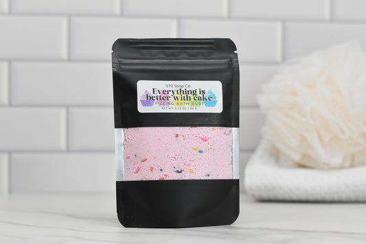 A black bag of 570 Soap Co.'s Birthday Cake Fizzing Bath Dust, adorned with the slogan "Everything is better with cake," rests on a white surface. In the background, against a white tiled wall, a soft bath sponge infused with moisturizing apricot kernel oil promises a sweet escape.