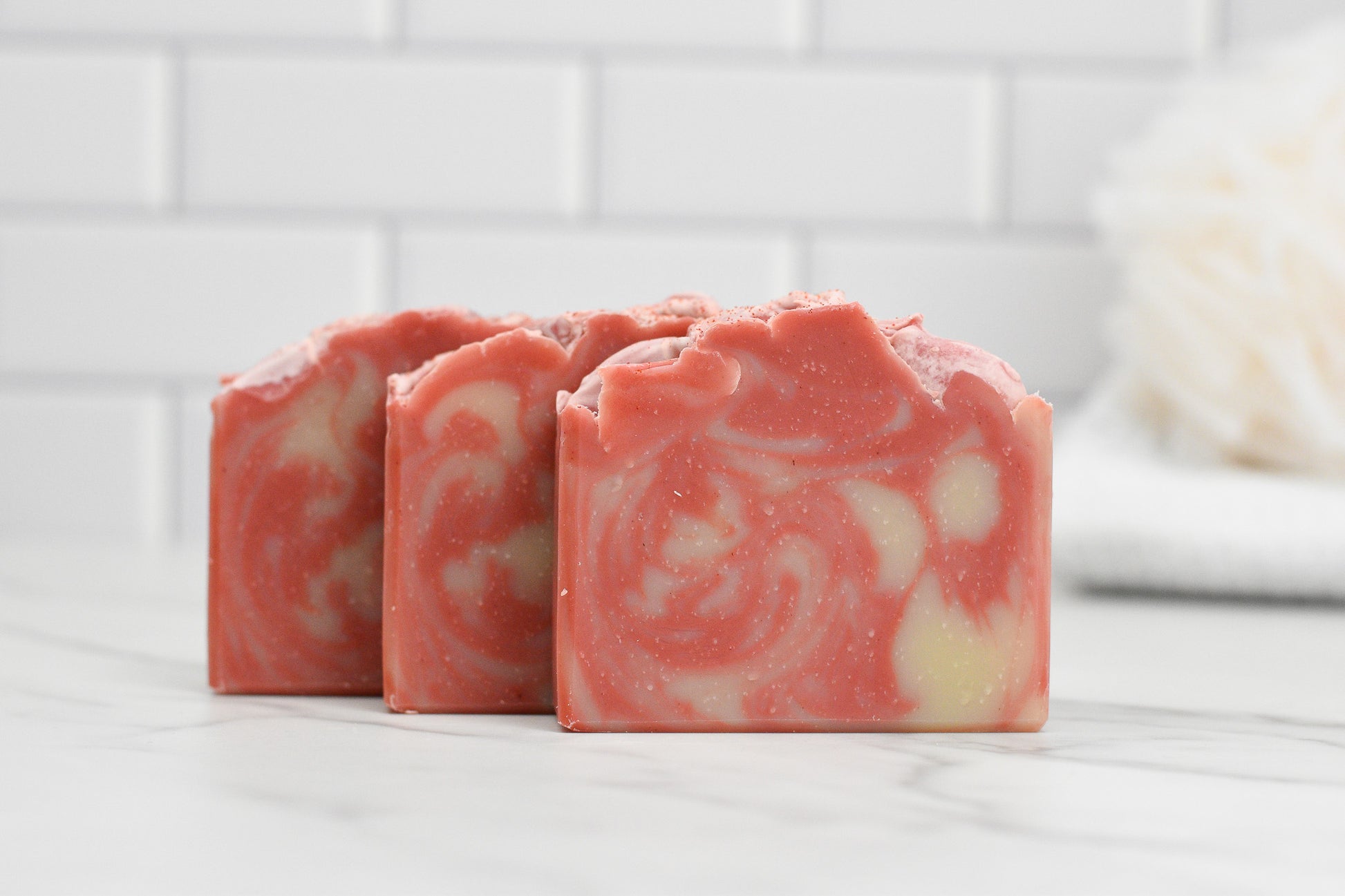 Three Candy Cane Lane Bar Soaps by 570 Soap Co., featuring pink and cream swirls, are elegantly arranged on a white marble surface. The setting includes white subway tiles in the backdrop, with a blurred bath puff accentuating the pristine ambiance. Indulge in its moisturizing formula for a refreshing cleanse.