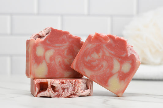 Two bars of 570 Soap Co.'s Candy Cane Lane Bar Soap are displayed against a white tiled background. One stands upright, while the other leans, highlighting its marbled design and twisted peppermint essence. A blurred bath sponge in the background hints at the soap's moisturizing formula.