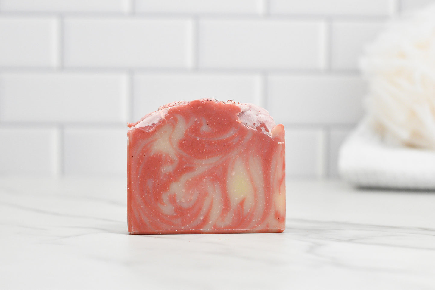 A bar of 570 Soap Co.'s Candy Cane Lane Bar Soap, featuring a pink and white marbled design, rests on a marble countertop. Its textured top holds a moisturizing formula. The background showcases a white subway tile wall with a soft, blurred white towel.