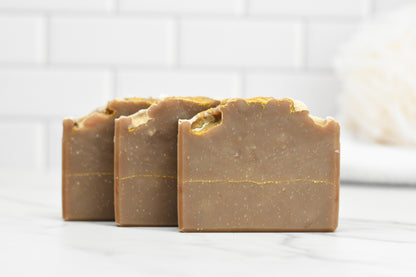 Three bars of 570 Soap Co.'s Frankincense & Myrrh Bar Soap, releasing ancient fragrances, are arranged against a white tile background. The soap showcases a natural brown shade and features a rustic, textured surface, with a bath sponge softly blurred in the background.