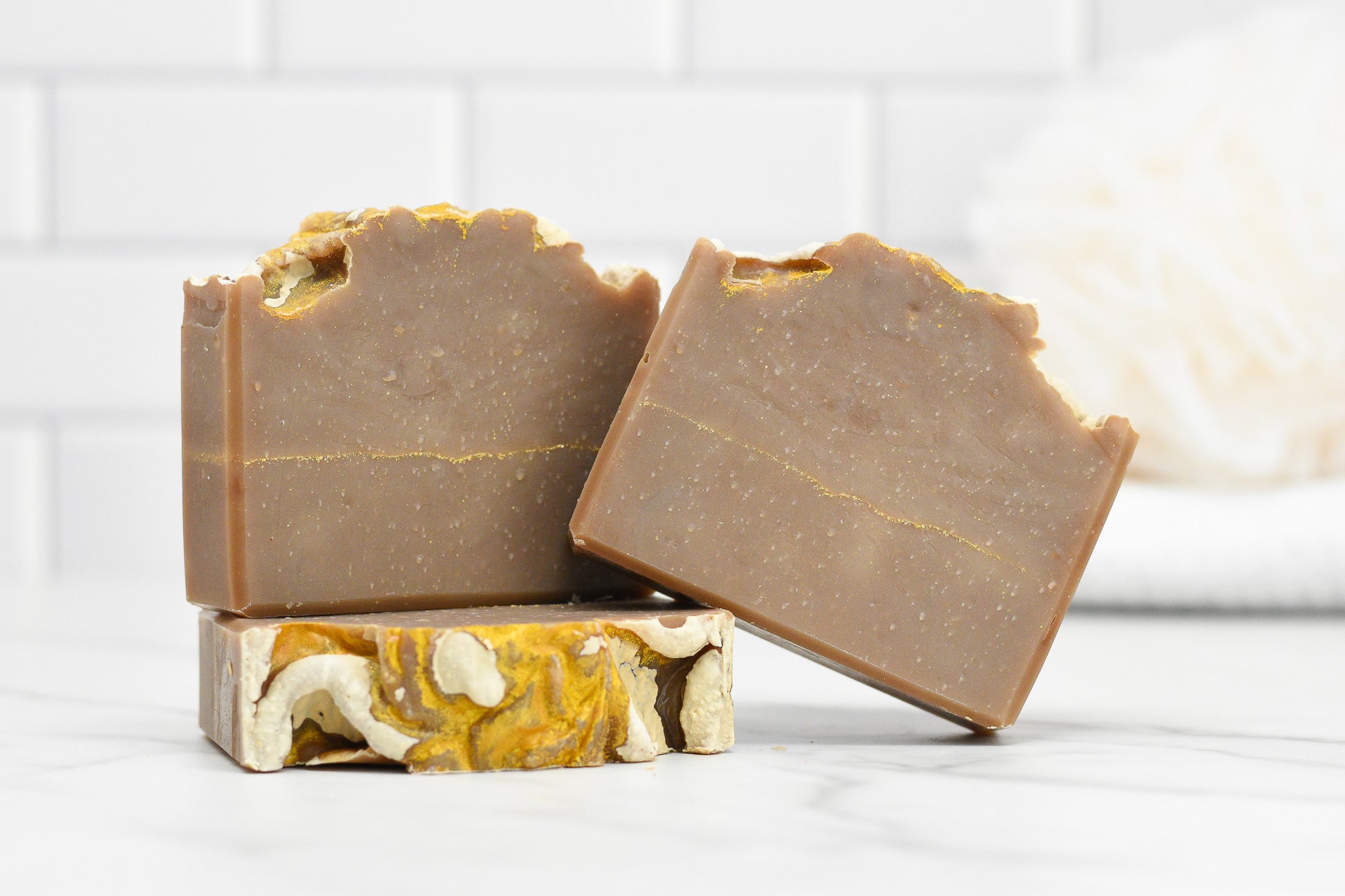 Three Frankincense & Myrrh Bar Soaps from 570 Soap Co. are stacked on a marble surface. Two bars lean against each other, highlighting their natural, textured brown finish with a subtle gold stripe. These exquisite creations embody artisanal craftsmanship. In the background, a blurred white sponge adds depth.