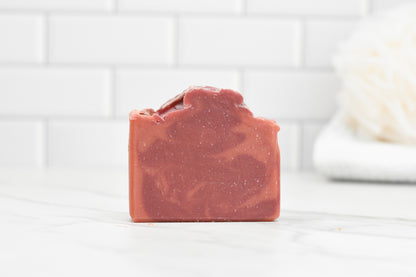 The Elf Sweat Bar Soap from 570 Soap Co., with its pinkish-red hue and rough natural texture, sits on a marble surface. The background features white subway tiles and an out-of-focus white object, possibly a towel, enhancing the enchanting scene.