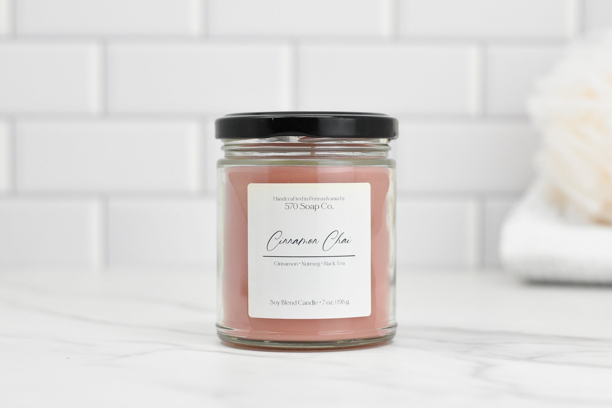 A Cinnamon Chai Soy Blend Candle by 570 Soap Co. rests on a marble countertop against white tiles, featuring pinkish-brown toxin-free wax and a sleek black lid.