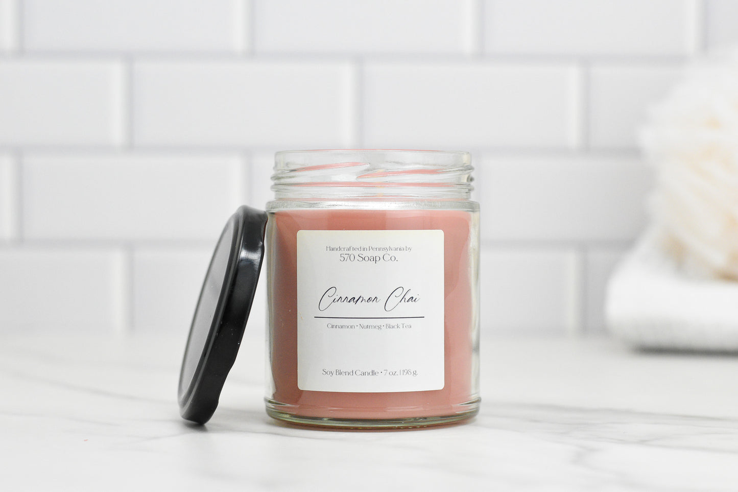 A 570 Soap Co. Cinnamon Chai soy blend candle with a black lid sits on a marble countertop. A white tile wall and blurred white cloth in the background emphasize its toxin-free wax composition.