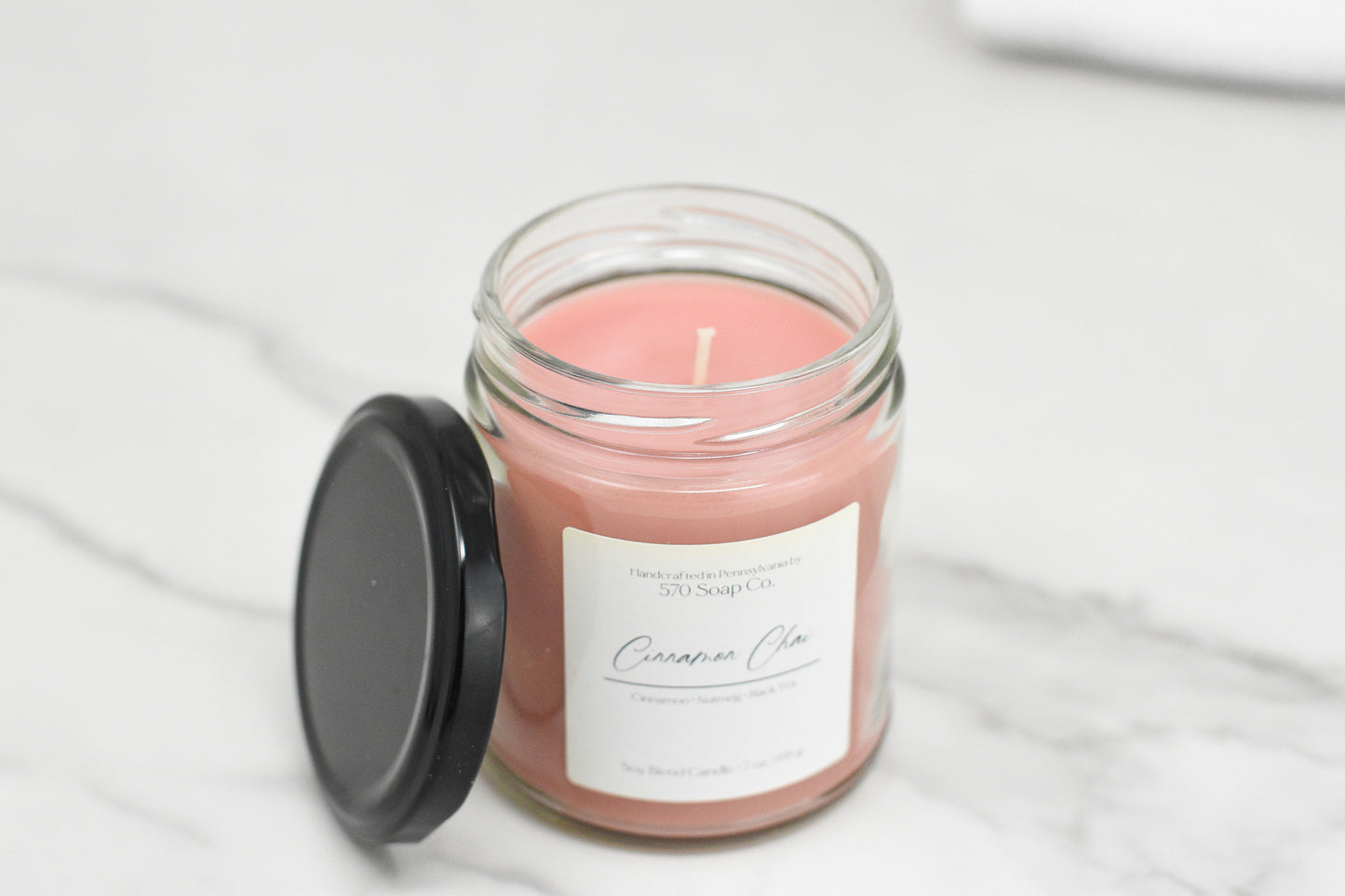 A pink Cinnamon Chai Soy Blend Candle by 570 Soap Co. rests elegantly on marble in a glass jar with a black lid. The label suggests cinnamon and nutmeg notes, while the toxin-free wax ensures a cleaner burn.