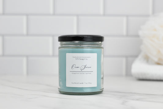 An Ocean Sunrise Soy Blend Candle from 570 Soap Co., featuring a black lid, is positioned on a white countertop against a tiled backsplash. The light blue wax emits a calming marine fragrance. In the background, a white loofah is partially visible, enhancing the peaceful ambiance of this candle arrangement.