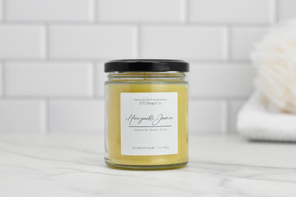 A Honeysuckle Jasmine Soy Blend Candle from 570 Soap Co., featuring a black lid, rests on marble. Its floral elegance enhances the scene with a white tiled wall, complemented by a soft-focus white bath scrub.