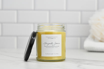 A yellow Honeysuckle Jasmine soy blend candle by 570 Soap Co. sits in a glass jar with a black lid on marble. The background features soft white tiles and a fluffy cream object, adding floral elegance.