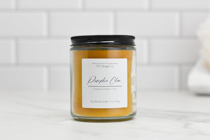 A Pumpkin Chai Soy Blend Candle from 570 Soap Co., housed in a glass jar with a black lid, emits an autumn aroma. The label features "Pumpkin Chai, Cardamom/Pumpkin/Clove" and indicates its weight as 7 oz./198 g. It is placed against a white tiled background for a cozy seasonal ambiance.