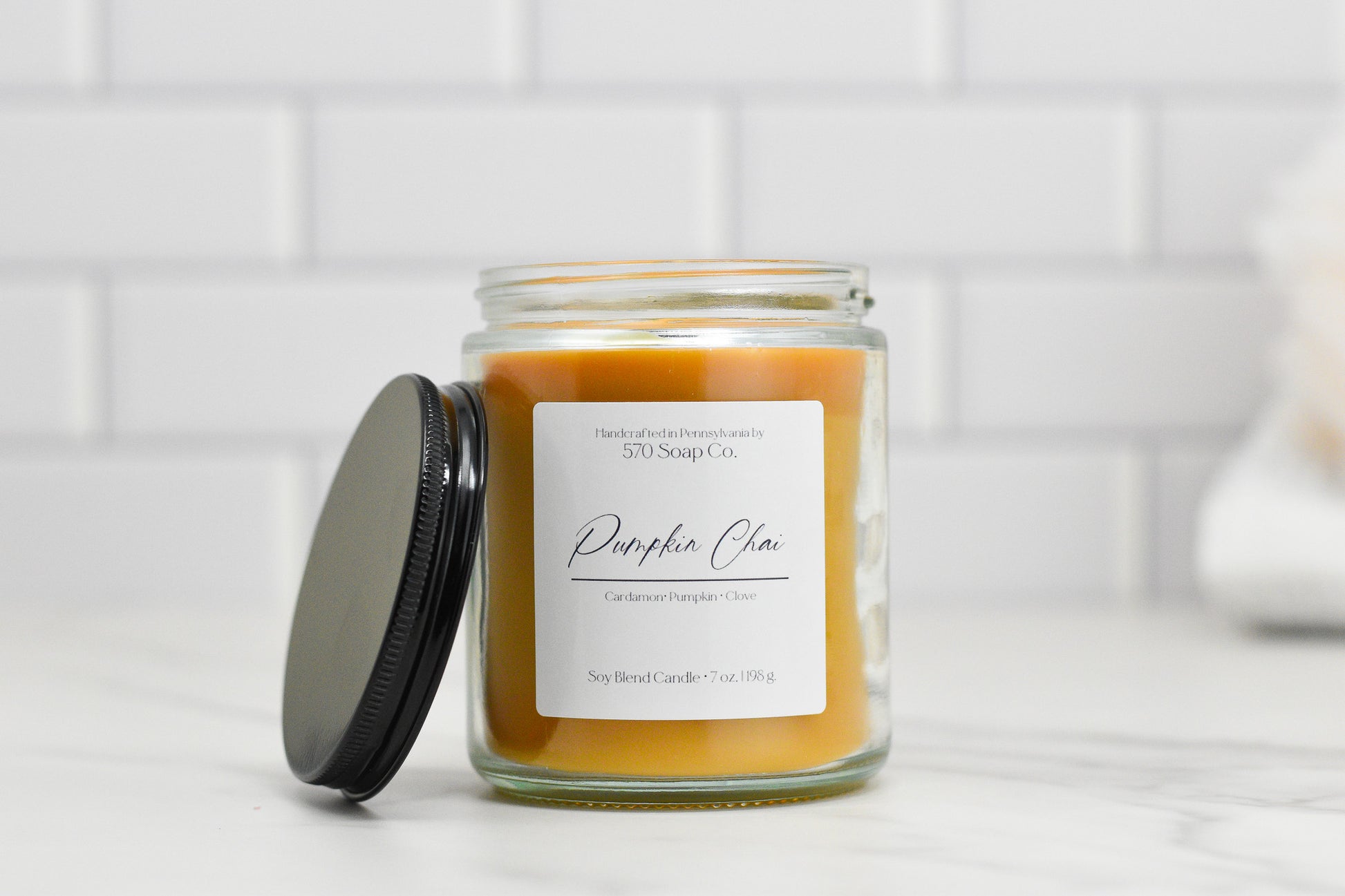 A glass jar of 570 Soap Co.'s Pumpkin Chai Soy Blend Candle, featuring a black lid and adorned with a label highlighting cardamom, pumpkin, and clove notes, rests elegantly on a white marble surface with a soft-focus white tile backdrop, embodying the quintessential autumn fragrance.