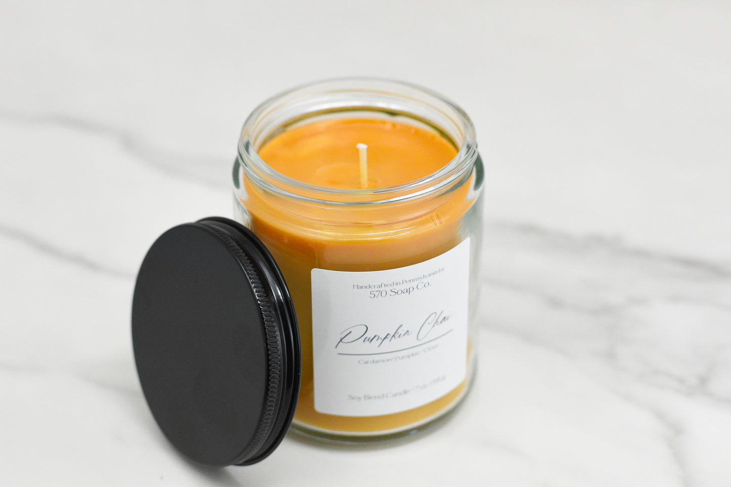 A Pumpkin Chai Soy Blend Candle from 570 Soap Co. is gracefully displayed on a marble surface, featuring orange wax and topped with a black lid. This candle embodies the quintessential autumn fragrance, making it ideal for inviting and cozy evenings.