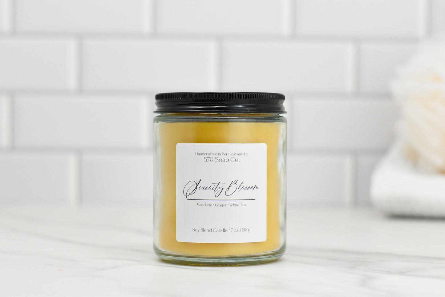 A Serenity Blossom Soy Blend Candle from 570 Soap Co. is showcased against a white tiled backdrop, featuring a black lid and a label that reads "Serenity Bloom, Mandarin Orange, White Tea." This candle is made of toxin-free soy blend wax with light orange coloring to create a calming atmosphere.