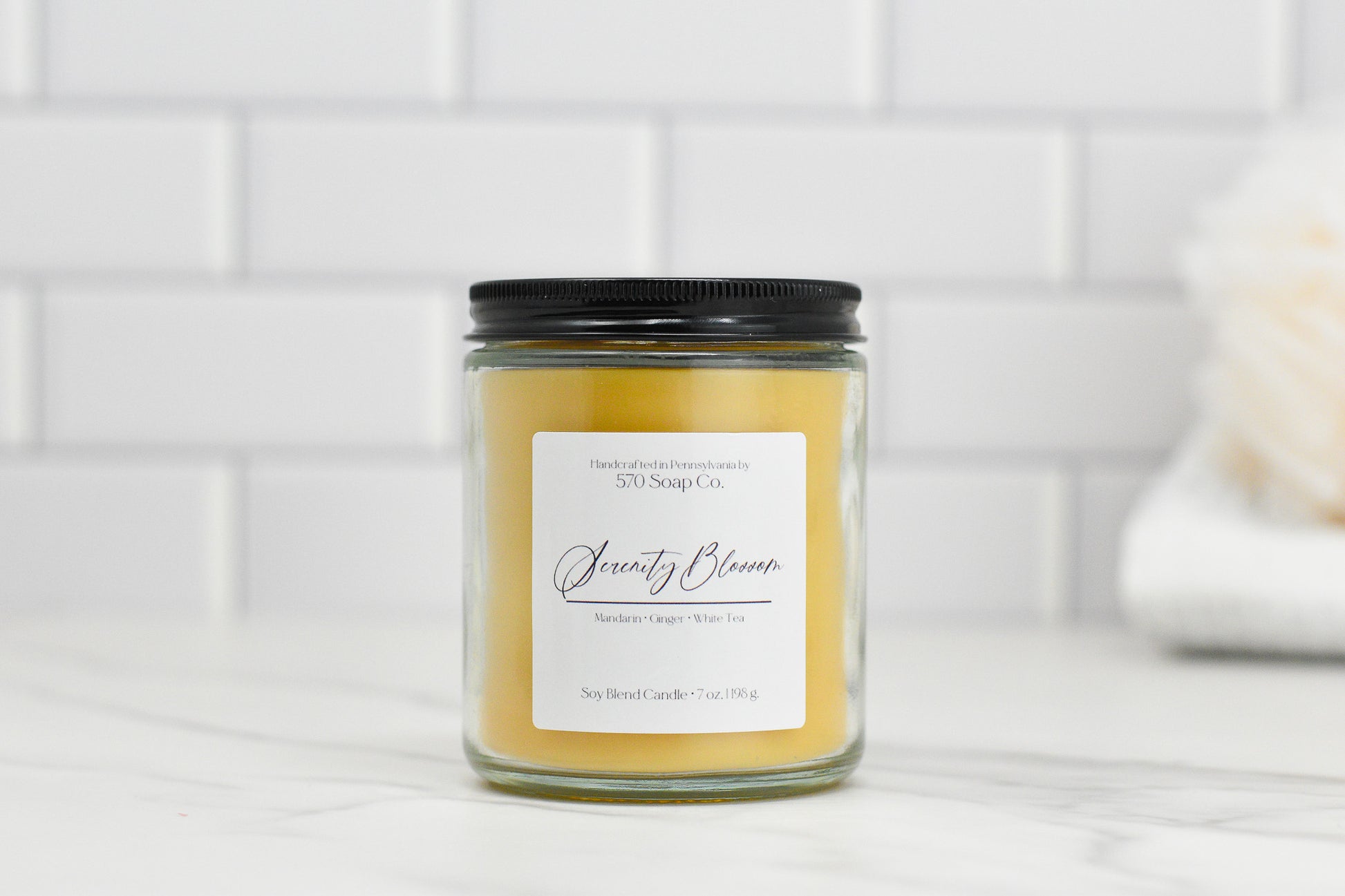 A Serenity Blossom Soy Blend Candle from 570 Soap Co. is showcased against a white tiled backdrop, featuring a black lid and a label that reads "Serenity Bloom, Mandarin Orange, White Tea." This candle is made of toxin-free soy blend wax with light orange coloring to create a calming atmosphere.