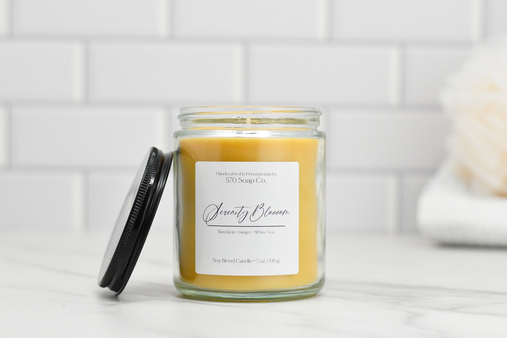 A jar candle, the Serenity Blossom Soy Blend Candle by 570 Soap Co., is on a counter with a black lid leaning against it. Crafted from toxin-free soy blend wax, it fills the air with fragrances of Mandarin, Ginger, and White Tea. In the background, there is a white tiled wall.