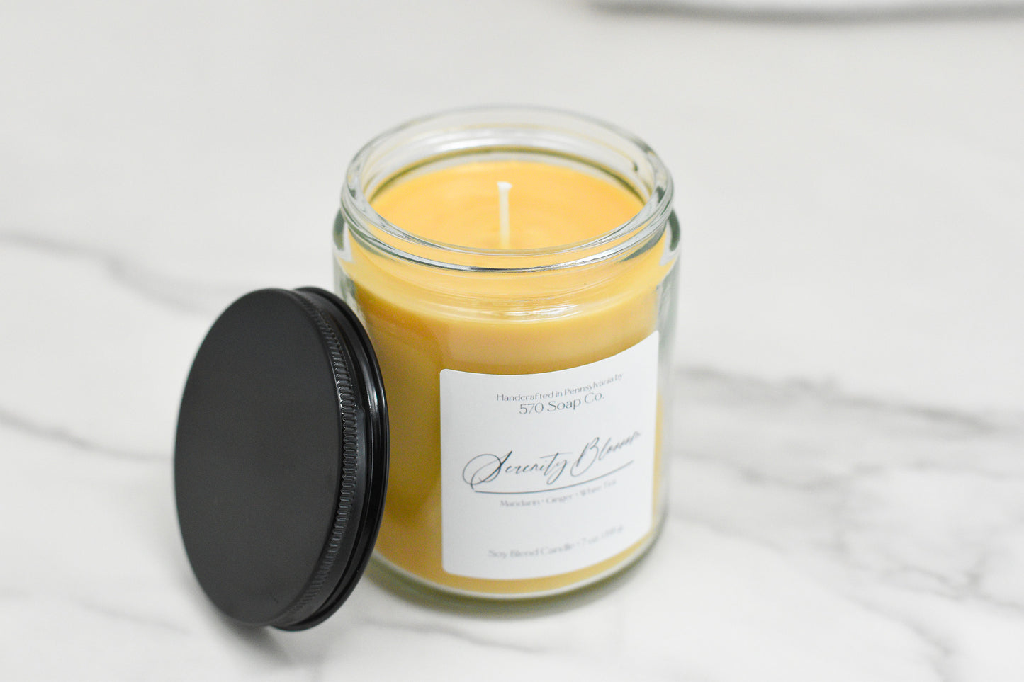 A Serenity Blossom Soy Blend Candle by 570 Soap Co. flickers gently in a clear glass jar with a black lid beside it on a marble surface, crafted from toxin-free soy blend wax. The label features decorative text that adds an elegant touch.