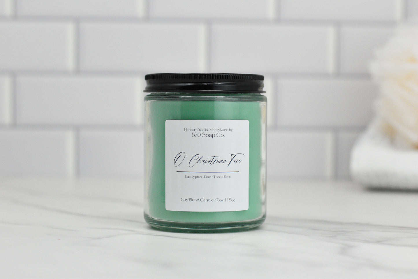 A green O Christmas Tree soy blend candle by 570 Soap Co. sits on a marble countertop against a white tiled wall, featuring Eucalyptus Pine and Terebinth scents for a winter forest aroma.